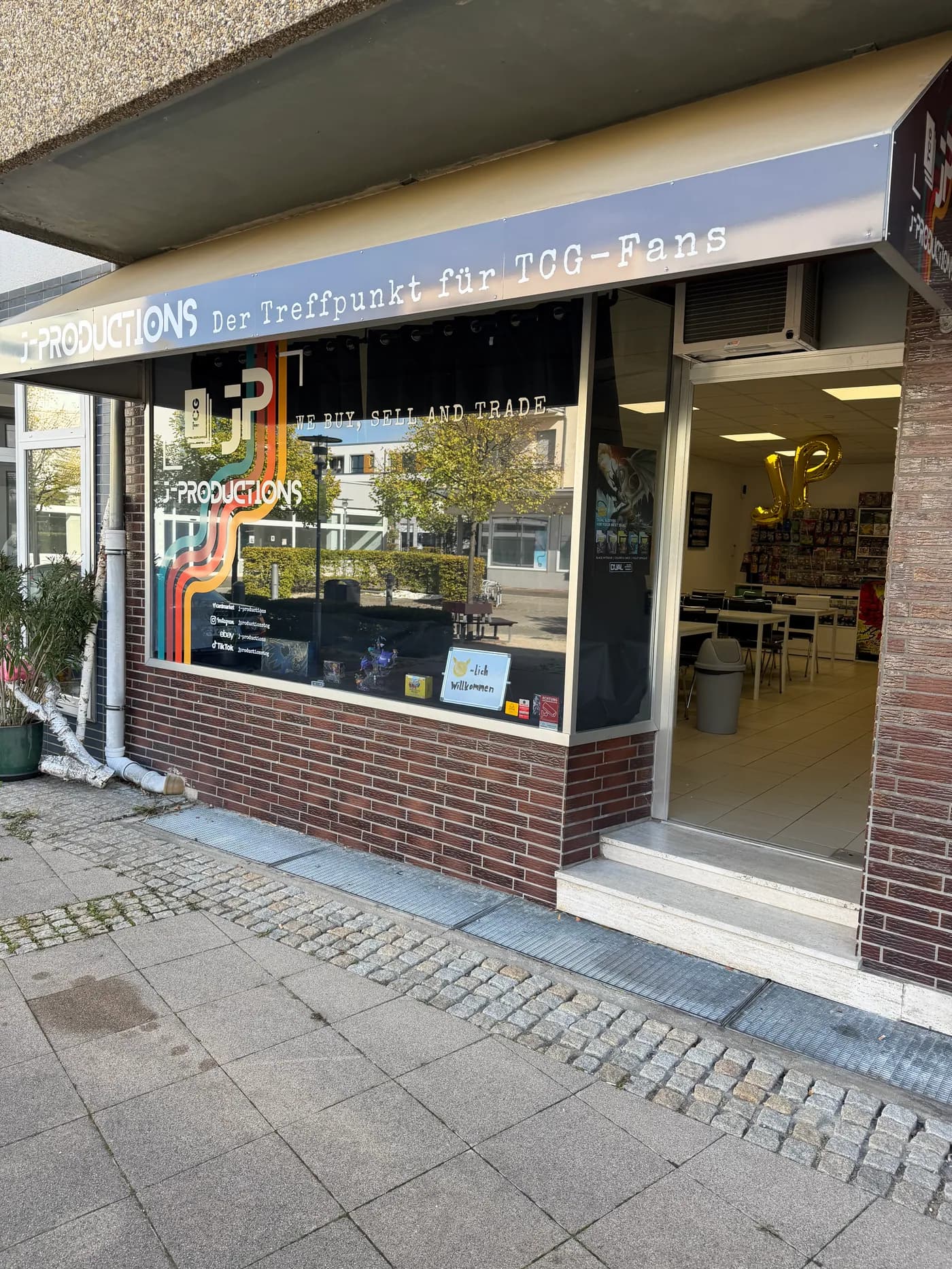 J-Productions von außen: Schaufenster mit Logo, Kartenpräsentation und geöffnetem Eingang